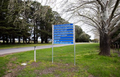 This image shows a blue sign situated on green grass with a road and a row of trees in the background. 
