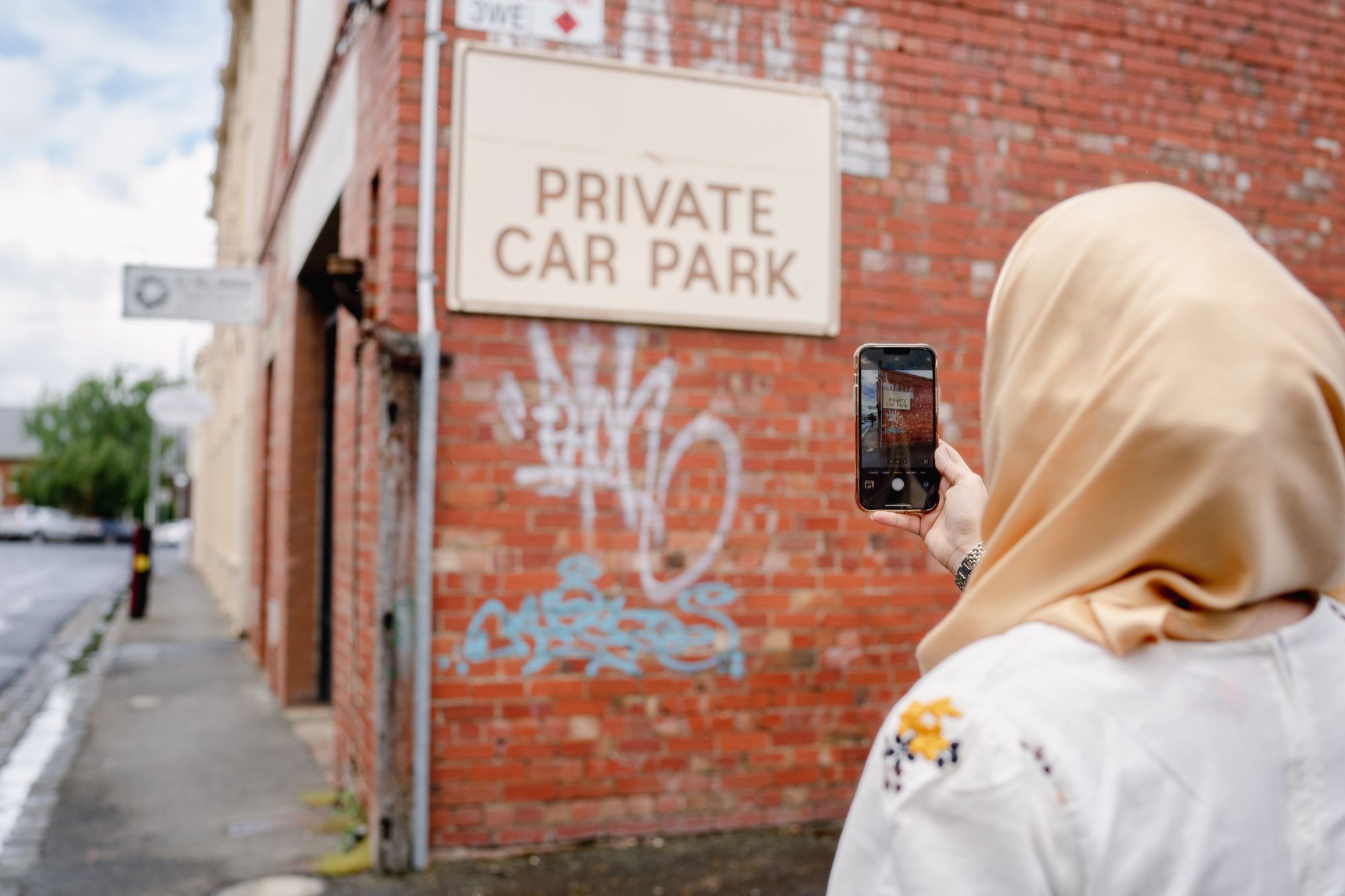 A woman in a headscarf takes a photo on her phone of graffiti on a red brick wall. 