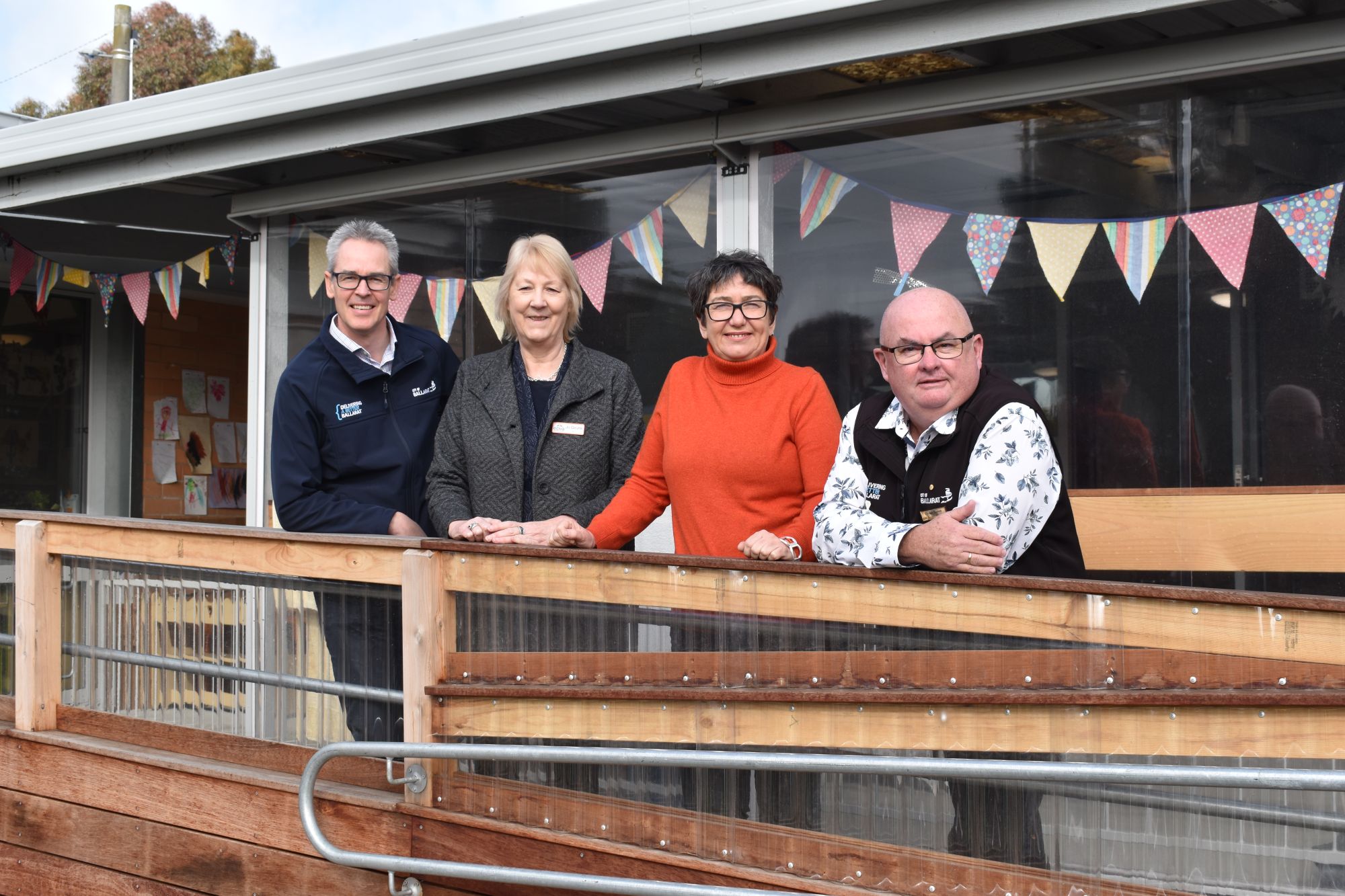 Four people, including two men and two women, stand on a ramp with bunting in the window behind them. 