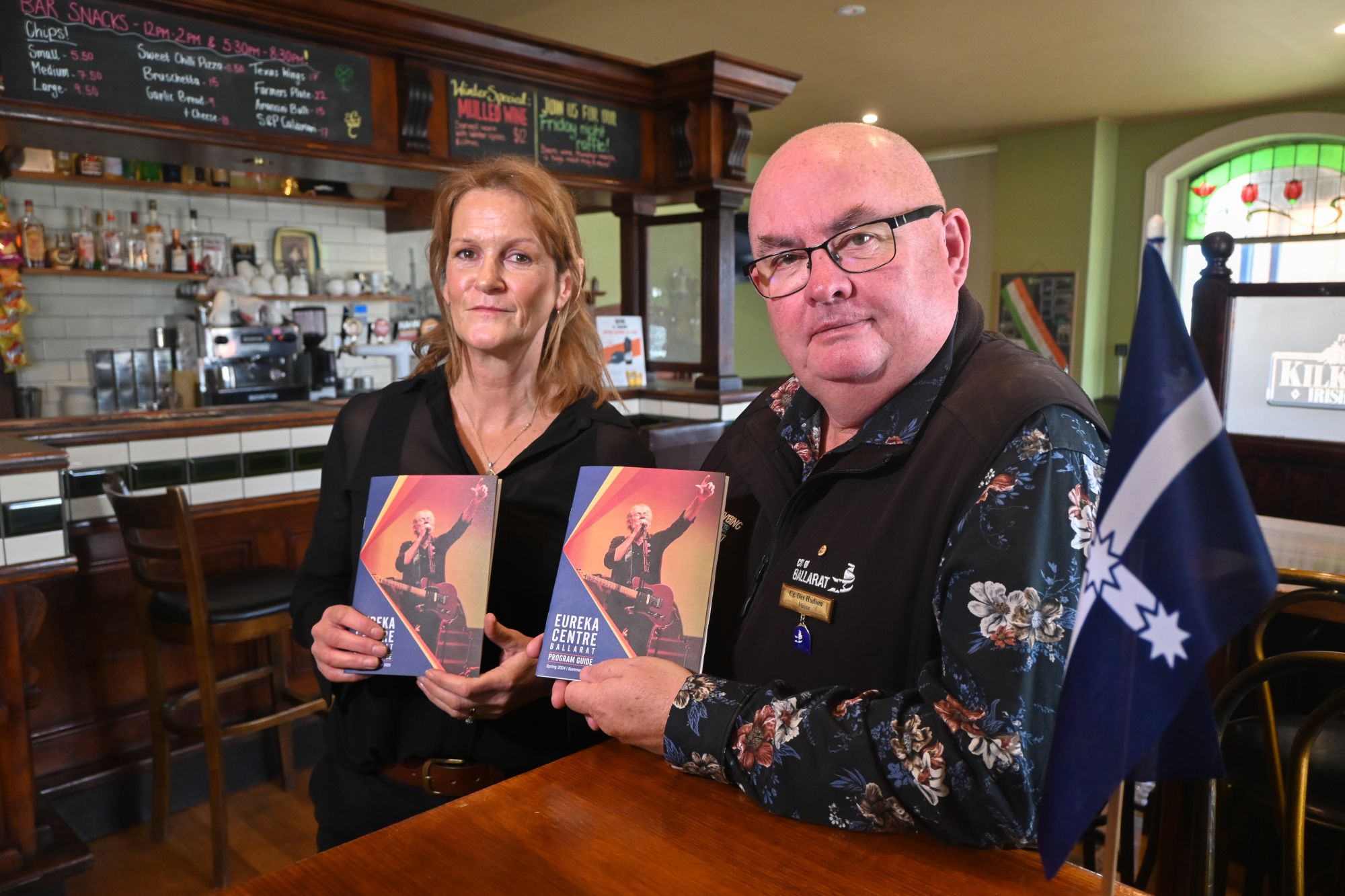 A man and woman stand in an Irish pub, holding two brochures, with a small Eureka flag on the side.