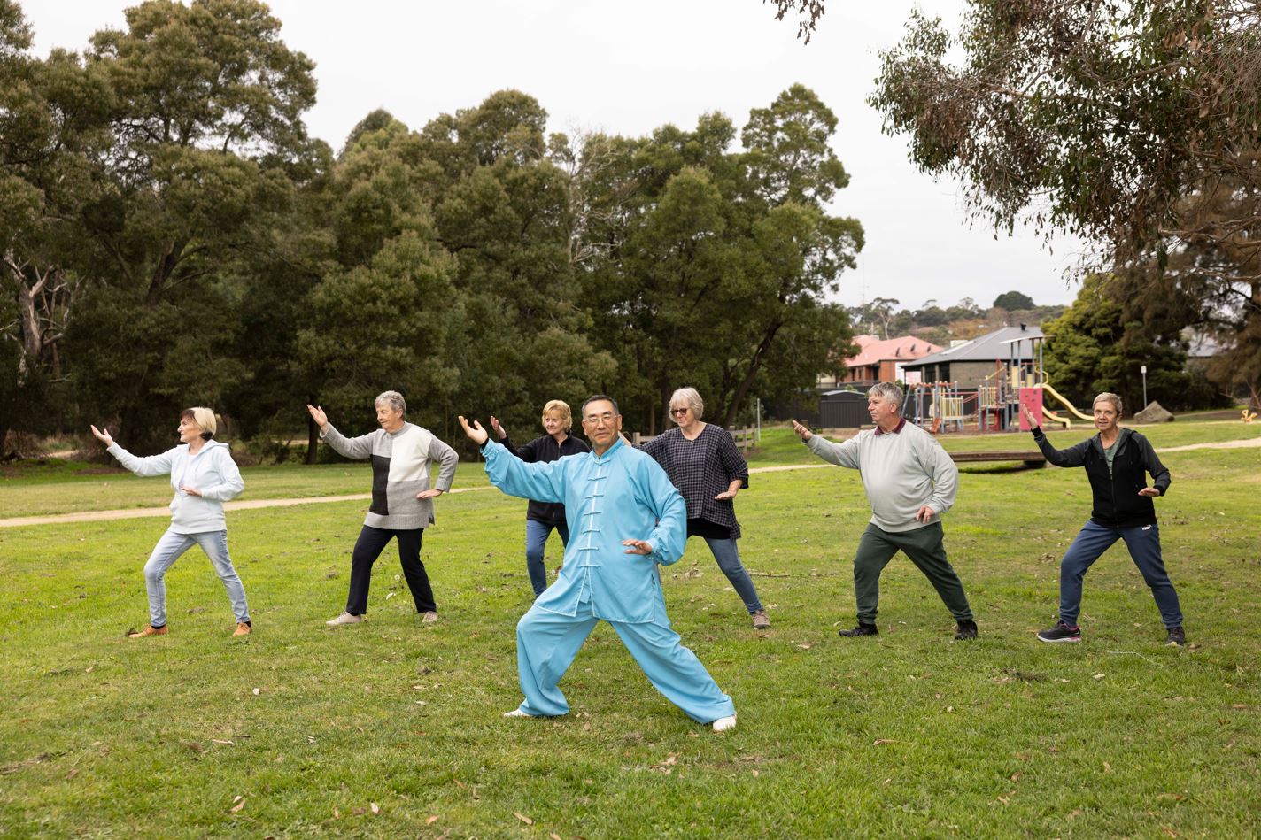 A group of people, with a man in a blue outfit in the front, do Tai Chi on the grass in front of a park. 