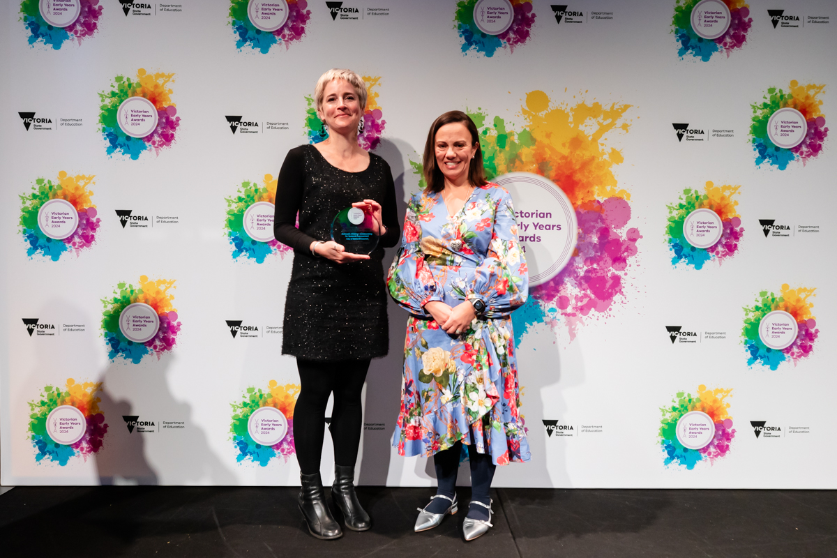 Two women stand in front of a white background with colourful logos on it. The women on the right holds a glass trophy. 