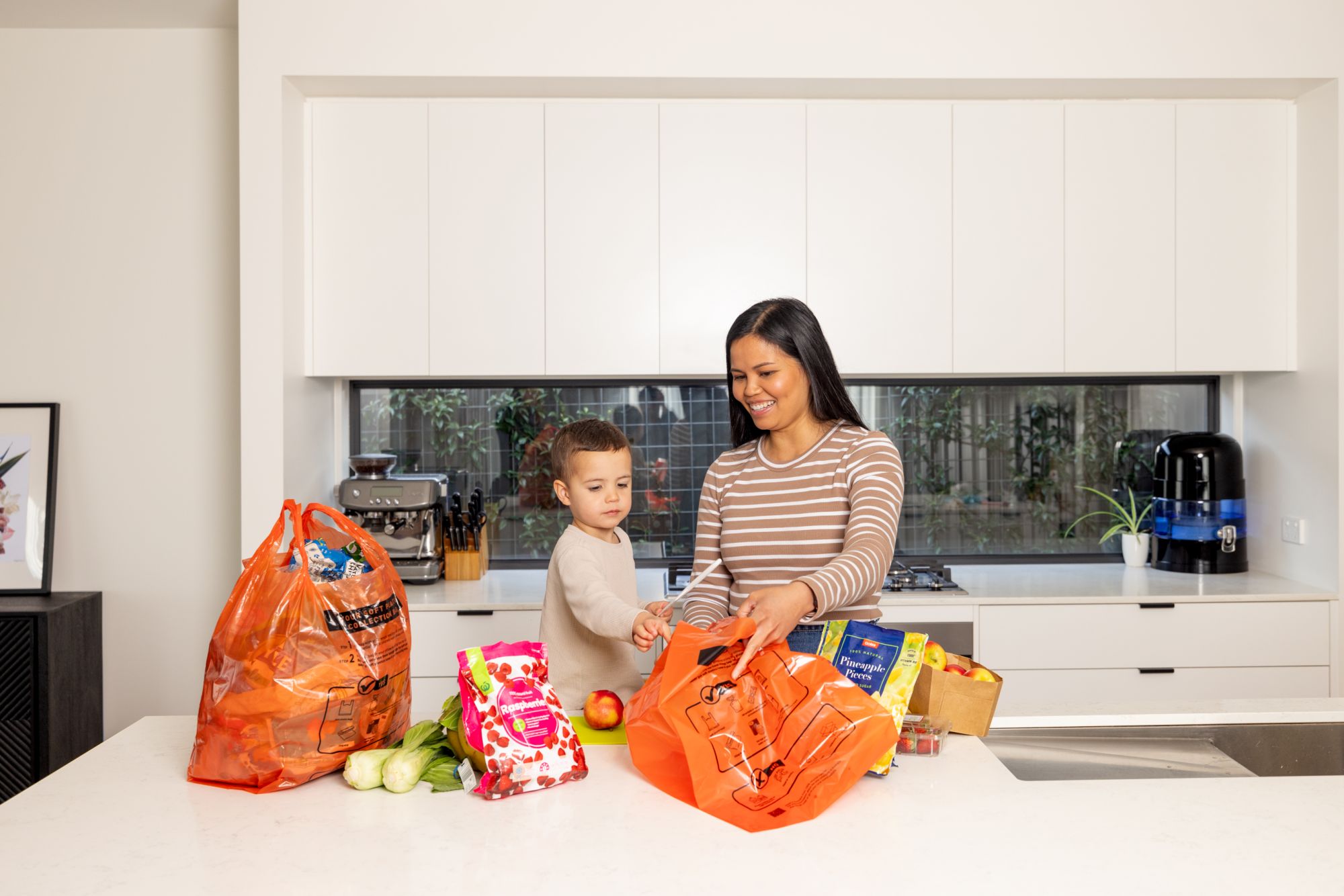 A woman and young boy stand at a kitchen bench with two large orange rubbish bags - the boy puts a piece of soft plastic rubbish into one of the orange bags. 