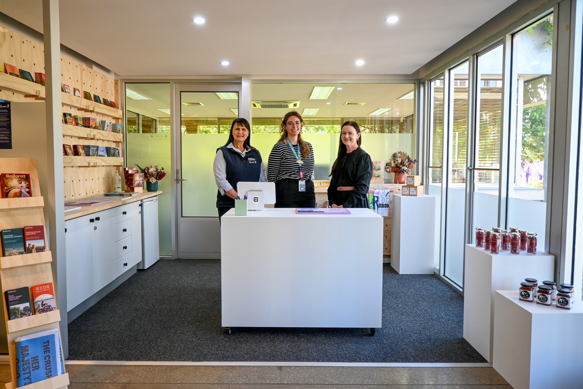 Three women stand behind a white desk in a light room with brochures on one wall. 