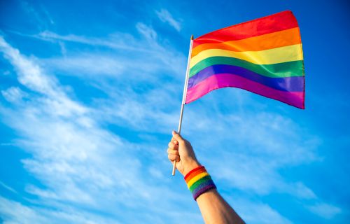 A hand with a rainbow wristband holds a rainbow flag against a blue sky