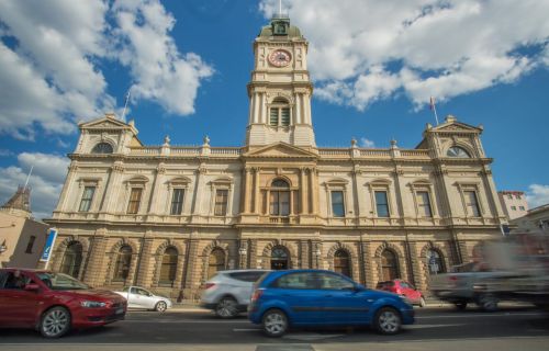 Ballarat Town Hall stands against a blue sky with some clouds, with some cars driving in front of it. 