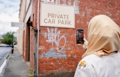 A woman in a headscarf takes a photo on her phone of graffiti on a red brick wall. 