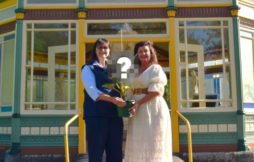 Two women stand in front of a rotunda holding a plant between them. The top of the plant has a question mark over it. 