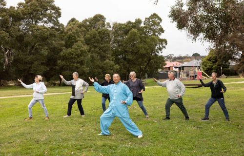 A group of people, with a man in a blue outfit in the front, do Tai Chi on the grass in front of a park. 