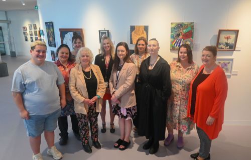 A group of people stand in front of a white wall covered with different pieces of art. 