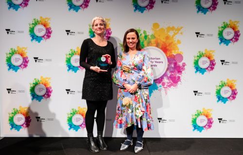 Two women stand in front of a white background with colourful logos on it. The women on the right holds a glass trophy. 