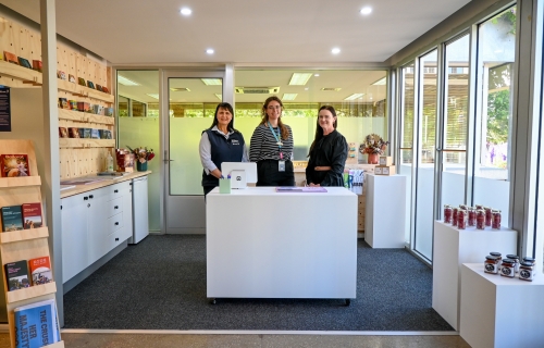 Three women stand behind a white desk in a light room with brochures on one wall. 