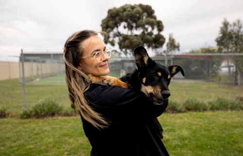 A woman holds a dog with green grass and fencing in the background. 