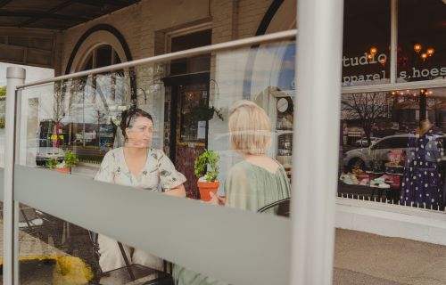 Two women sit in the outdoor dining area of a café, behind glass panels