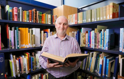 Information Services Librarian Simon Jacks holding book and standing infront of book case