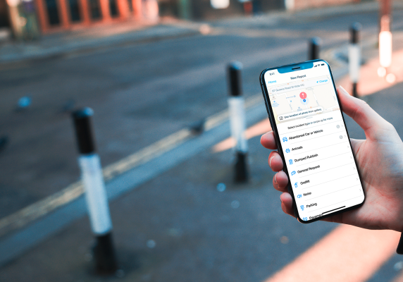 A hand holds a mobile phone with the Snap Send Solve app open. An empty urban road is in the background. 