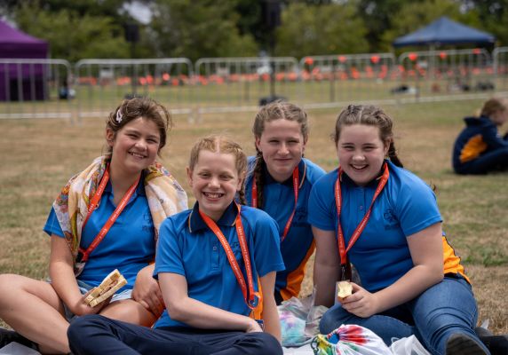 Four girls in blue shirts and orange lanyards sit on a grassed area. 