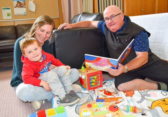 On the left, a young boy sits in a woman's lap and on the right, City of Ballarat Mayor Cr Des Hudson is seated on the ground and holds an open picture storybook. 