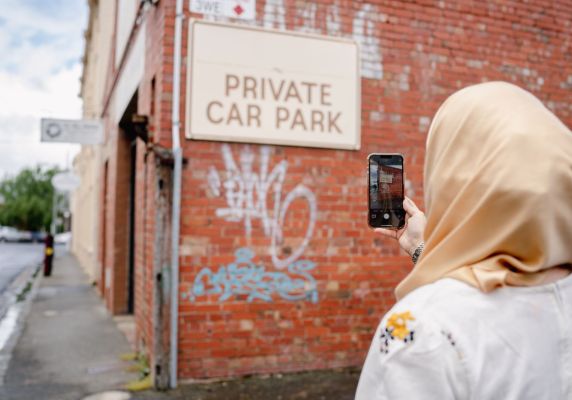 A woman in a headscarf takes a photo on her phone of graffiti on a red brick wall. 