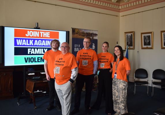 Three men and two women wearing bright orange T-shirts stand in front of a screen that says 'Join the Walk Against Family Violence'