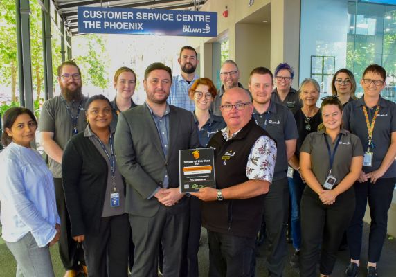 A group of people stand in front of the City of Ballarat Customer Service Centre. The front two people are holding a framed certificate. 