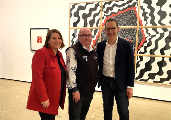 A woman in a red jacket and two men stand in front of a black and white geometrical artwork. 