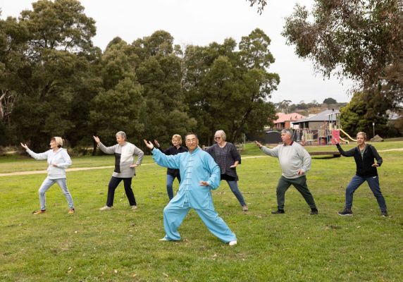 A group of people, with a man in a blue outfit in the front, do Tai Chi on the grass in front of a park. 