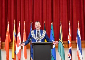 Current City of Ballarat Mayor Cr Daniel Moloney is pictured wearing the Ballarat Mayoral Chains in front of a red curtain and a row of flags.