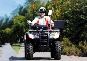 A man a in a helmet drives a large quad bike on a tree-lined footpath. The quad bike has a laptop and camera equipment attached to the front. 