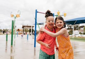 A girl in an orange jumpsuit (left) hugs a girl in a red shirt (right) with splash park equipment in the background. 