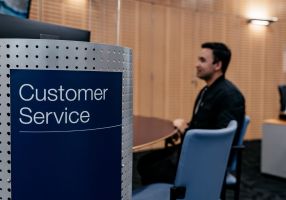 In the foreground, there is a dark blue sign with 'Customer Service' written on it. In the background is a man with sitting at a desk with a blue chair next to him. 